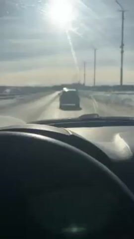 The video shows a view from inside a car, looking out the windshield at a road on a sunny day. The road is mostly clear, but there is snow on either side. There are a few other cars on the road, and the car in front is the closest. There are also street lights on either side of the road. The steering wheel and dashboard of the car are visible in the foreground. The steering wheel has a Skoda logo on it.