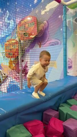 A young child wearing a light-colored t-shirt and shorts jumps off a blue padded platform into a ball pit filled with pink and purple cubes. The child is mid-air, appearing to be falling or landing. Behind the child, a net encloses the play area, and a colorful mural with abstract shapes and a digital scoreboard is visible on the wall. The child is the only person in the frame.