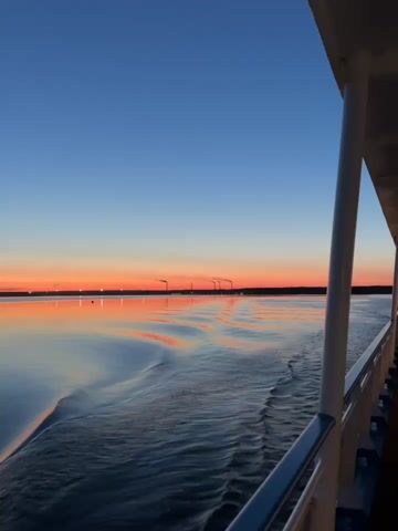 A ship sails on calm water during sunset. The sky is a gradient of deep blue to orange. The water reflects the colors of the sky, with gentle waves creating patterns. In the distance, a faint silhouette of industrial chimneys can be seen against the horizon. The railing of the ship is visible in the foreground, indicating the perspective of someone on board.