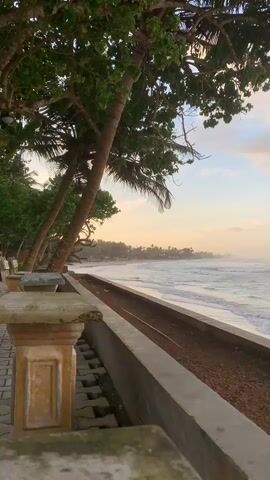 The video shows a serene beach scene at Kathaluwa, Sri Lanka. The time of day appears to be late afternoon or early evening, with a warm, soft light casting a gentle glow over the landscape. The sky is a mix of pale blue and soft orange hues, with scattered clouds, suggesting fair weather.

The foreground features a stone balustrade with decorative pillars and a tiled walkway. Beyond this, a concrete retaining wall runs parallel to the beach, with a strip of dark soil behind it, possibly indicating a garden area or a construction zone. Several palm trees with lush green foliage dominate the left side of the frame, their trunks leaning towards the sea, creating a picturesque canopy. Two park benches are visible under the trees, offering a place to rest and enjoy the view.

The main subject is the ocean, with gentle waves breaking on a sandy beach. The water is a mix of blue and white, reflecting the soft light of the sky. In the distance, the coastline curves, with a line of buildings and more palm trees visible, suggesting a developed area with resorts or houses. The overall mood is peaceful and tranquil, evoking a sense of relaxation and natural beauty. No people are visible in the frame. There is no visible text in the media file.