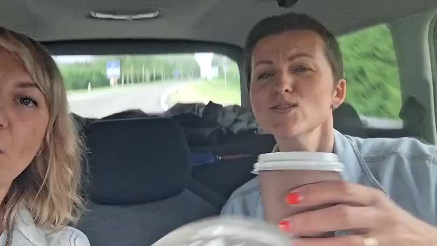 The video shows two women in the back seat of a car. The woman on the left has blonde hair and is wearing a white shirt. The woman on the right has short brown hair and is wearing a light blue jacket over a purple shirt. She is holding a white cup and a cell phone. The car appears to be moving, as the background is blurred. The women are both smiling and looking at the camera. The woman on the right is speaking. The scene is consistent with a casual car ride.
