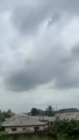 The video presents a static shot of a residential area under a heavily clouded, overcast sky. Several single-story houses with weathered roofing are visible amidst green foliage, suggesting a suburban or semi-urban setting. A tall telecommunications tower rises in the background. The angle of the shot implies it was taken from an elevated position, possibly a higher floor within one of the buildings.