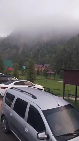 The video shows a parking lot with several cars parked. In the background, there are mountains covered in fog, trees, and some houses. The weather appears to be overcast or foggy. The cars are of various types and colors, including a silver SUV in the foreground, a white car behind it, and a black truck further back. The parking lot surface is dirt. There are also some trees and a fence visible.