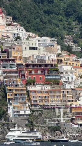 The video shows a view of a hillside town on a sunny day, with boats in the water below.  The town is densely built with houses of various colors, including white, yellow, and red.  Many of the houses have balconies, and there is some vegetation on the hillside among the buildings.  The water is relatively calm. There are several boats in the water, including a larger white boat and some smaller boats. There are people visible on the shore near the boats.