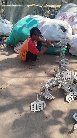 The video shows a young person sitting on the ground in an outdoor setting, surrounded by large sacks and piles of white plastic objects. The person is wearing a red and orange shirt and black pants and is holding a metal rod connected to some of the plastic pieces. The plastic objects appear to be some kind of disassembled or discarded component, possibly from a fan or a similar device, as they have a grid-like structure with circular holes.

The person is actively engaged with the plastic pieces, suggesting they might be sorting, cleaning, or dismantling them. The overall scene appears to be a recycling or waste processing area, given the presence of the large sacks filled with materials and the scattered components. The ground is dusty and uneven, typical of an outdoor industrial or informal recycling site.

Sunlight is casting shadows on the ground, indicating it is daytime, likely a sunny day. The person's posture and focus on the task suggest concentration rather than distress. In the background, there are large white sacks, one of which has a graphic of a person's face on it. There are also some glimpses of what might be bicycles or motorbikes, hinting at a bustling environment. The location context provided, Kertagena Laok, Indonesia, further reinforces the idea of a potentially informal or developing area where such waste processing activities might occur. No discernible text is legible on the plastic items themselves, but the graphic on the sack is visible.