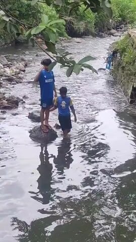 Two boys are in a shallow river. One boy stands on a rock wearing a blue "Allstar" shirt, blue shorts, and no shoes. The second boy stands in the water wearing a blue and yellow shirt, dark shorts, and no shoes. The river is rocky with green foliage on either bank. The water is murky but clear enough to show reflections. The boy in the water gestures with his right hand and drinks with the left. The other boy is still.