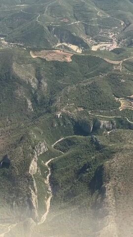 This is an aerial, high-angle shot of a mountainous, forested landscape under a bright sky. The terrain is characterized by steep slopes, deep valleys, and winding roads that snake through the greenery. Terraced fields are visible on some hillsides, indicating human agricultural activity. In the distance, a small cluster of buildings, suggesting a village or town, is nestled in a valley. The overall impression is one of rugged natural beauty with evidence of human presence in a remote, hilly region.