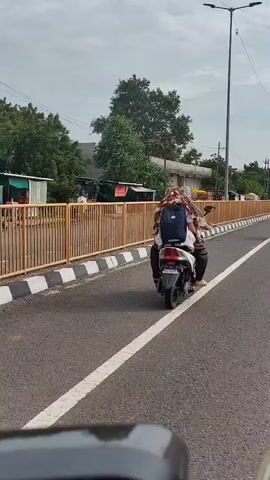 Two people are riding on a white scooter on a paved road. The person in front is wearing a white shirt and the person behind is wearing a patterned headscarf and carrying a black backpack. They are riding on the right side of the road, which has a white line marking the lane. A yellow metal fence runs along the left side of the road. Trees and buildings are visible in the background under a cloudy sky. The video appears to be recorded from another vehicle.