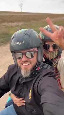 Two people are seen riding a quad bike on a dirt track in an arid, open landscape, likely Lalla Takarkoust, Morocco. The video is a selfie taken from the perspective of the front rider, a man who is smiling broadly at the camera. He wears a black helmet with colorful accents, sunglasses, a red and white patterned bandana, and a black jacket. The passenger behind him, a woman, is also smiling (implied by her body language) and gives a peace sign with her right hand. She wears a green helmet, sunglasses, a multi-colored patterned scarf covering her lower face, and a light-colored top. Both helmets bear the brand name "VEGA". In the background, other quad bikes can be seen on the track, and a lone tree stands on a distant hill under an overcast sky. The scene depicts an enjoyable outdoor activity.