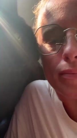 The video is a close-up selfie of a person wearing glasses, with a black cat positioned to their left. The cat is on or near the person's shoulder and is leaning in towards the person's face. The person is looking towards the camera, and the cat's head is partially obscuring the person's face. Sunlight is visible, creating lens flares and illuminating parts of the scene, particularly the person's face and hair. The cat appears to be interacting with the person's face, possibly nuzzling or nipping. The person is wearing a light-colored, possibly white, crew-neck top. The background is blurred and appears to be an indoor setting.