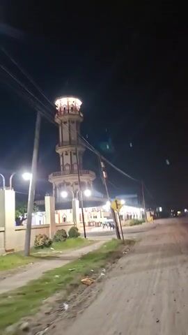 A nighttime video captures a tall, multi-tiered tower structure illuminated by lights. The tower has ornate balconies and decorative elements. In the foreground, a dirt road is visible, with a grassy shoulder and a lamppost. A yellow directional sign with arrows is mounted on a pole. Several people are seen in the background near the base of the tower and in a building adjacent to it. Overhead power lines stretch across the frame, and small, white specks, possibly insects or debris, are visible in the dark sky. The overall scene suggests a public place or building, possibly a religious site or a landmark, with activity occurring at night.