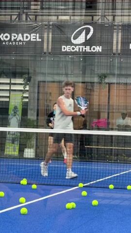 A young male padel player, wearing a white sleeveless shirt and patterned shorts, is on a blue court with a net in the foreground. He is in a crouched position, reaching down to pick up a yellow padel ball. Scattered across the court are numerous yellow padel balls. In the background, behind the net, a young female padel player, dressed in black, holds a racket. Above them, a dark banner displays logos for "Niko Padel Academy" and "Decathlon." The environment appears to be an indoor padel court with glass walls and a roof. The player's action suggests he is participating in a drill or practice session focused on collecting balls.