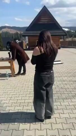 The video shows a behind-the-scenes look at a video shoot. A woman with long brown hair is standing in the foreground, facing away from the camera. She's wearing a black off-the-shoulder top and gray pants. In the background, a man wearing a long brown coat and a hat is leaning over a wooden table, adjusting a camera. The table has various items on it, including what appear to be lenses, a bottle of water, and other film equipment. The woman then sits on the table to be filmed. A black puffer jacket lies on the table in front of her. The setting is an outdoor paved area with a log cabin in the background and a view of forested hills. The sky is blue with some clouds.