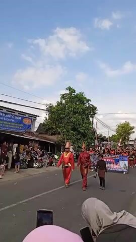 A low-angle shot captures a cultural parade moving down a street. Two men, dressed in matching red and gold traditional attire with conical hats, lead the procession, followed by a young boy in a similar outfit. Behind them, a banner reads "KARNAVAL BUDAYA RT 03 RW 02 NUSANTARA PULAU SUMATRA". A crowd of onlookers lines the side of the road, many using their phones to record the event. A "PLASTIC SHOP" sign is visible on a building in the background. The sky is a clear blue with scattered clouds. There are motorcycles parked on the side of the road.