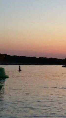 The video shows a body of water during sunset. In the foreground on the left, part of a green inflatable object with a red mushroom-shaped top is visible. The water has gentle ripples. In the middle distance, a person is standing on a paddleboard. Further out, a sailboat is visible, and then a small inflatable boat with at least two people in it. There are also several small buoys scattered across the water. The background consists of a dark treeline silhouetted against a sky with a gradient of colors from orange to pink to a light blue at the top. The overall atmosphere is calm and serene.