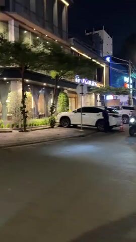 This video captures a street scene at night with several vehicles present. In the foreground, a street or sidewalk is visible, with shadows cast by overhead lights. To the left, a white SUV is parked, with its front bumper and headlights illuminated. Moving to the right, a black SUV is also parked, and a person wearing a dark cap and dark clothing is standing between the white and black SUVs, facing away from the camera, with their arms crossed. Further to the right, another white SUV is parked, with its rear visible. In the background, a modern building with large windows and lights is illuminated. Trees with green foliage are planted along the building's facade. A black fence with vertical slats and decorative plants attached is also visible in the background. The overall impression is that of an urban environment at night, possibly outside a restaurant or commercial establishment.