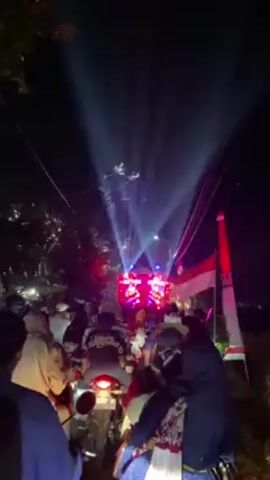 At night, a crowd of people on motorbikes are seen approaching a large, brightly lit structure on a narrow road lined with trees. The structure appears to be some sort of sound or lighting system, emitting beams of light that reach high into the sky. The lights change color from blue to white to red to green as the motorbikes draw closer. The Indonesian flag is visible on the right side of the road. Many individuals in the crowd are wearing face masks, and the overall atmosphere seems festive and lively.