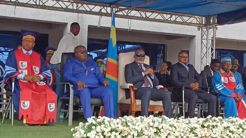 A group of people are seated in chairs on a raised stage, adorned with rows of white flowers in front of them. The people are wearing a variety of clothing including graduation gowns, suits, and casual attire. The flag of the Democratic Republic of the Congo is situated behind them, between two chairs. The environment appears to be outdoors.