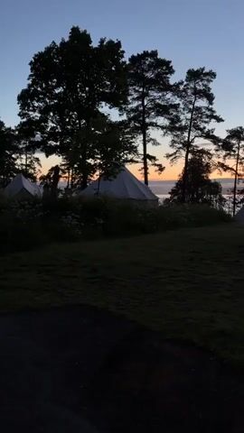 A person walks from the left to the right side of the frame in an outdoor setting at dusk. The person is silhouetted against a vibrant orange and blue sky, with a body of water in the background. Several tent-like structures are visible in the distance, interspersed among silhouetted trees. The foreground is a dark grassy area, with a narrow path at the bottom of the frame.
