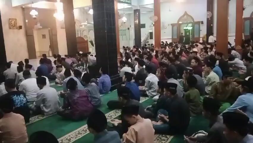 A video shows a crowded mosque interior with many men and boys sitting on a green carpeted floor. The men are dressed in various traditional Islamic clothing, including peci hats and sarongs. Some are interacting with each other, while others are looking towards the front of the mosque. The mosque features tall columns, chandeliers, and an arched doorway in the background. A person in a white shirt and dark pants is walking through the crowd. The lighting suggests it is daytime or evening within the mosque.