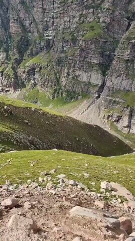 The video pans across a mountainous landscape. In the foreground, there is rocky, dry ground with scattered small stones and some sparse green vegetation. The middle ground features a steep, grassy slope with some terracing and scattered rocks. Further back, a dramatic, sheer cliff face with visible rock layers dominates the view. The background reveals a deep valley with more green slopes and distant, hazy mountains under a partly cloudy blue sky. The camera movement is a slow, upward and rightward pan.