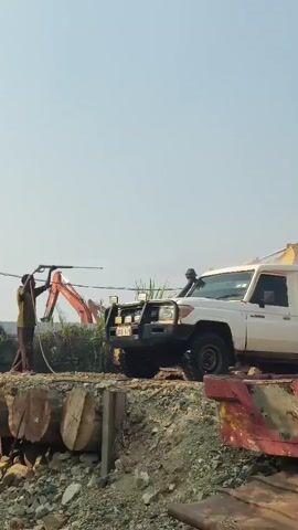 A person holding a long pole or pipe is positioned to the left of a white SUV. The SUV is parked on a dirt embankment next to a large, rusty metal object, possibly a barge. In the background, an orange excavator is visible. The sky is clear and bright.