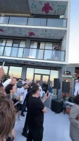 This video captures an outdoor event, likely a performance or concert, taking place in Lisi, Georgia. The scene is set on a rooftop or elevated platform with a clear blue sky overhead, suggesting daytime or early evening. A modern building with multiple balconies and large windows forms a backdrop, with "LISI EVENT HALL" visibly emblazoned on its facade.

Several people are present, both on a stage and in the audience. On the stage, there are at least three individuals who appear to be performers. One is wearing a white t-shirt, a red cap, and white pants. Another is in a white t-shirt and baggy white pants. A third individual, wearing a white t-shirt, dreadlocks, and a black beanie, is actively engaged in what seems to be a performance. He is wearing white pants and appears to be dancing or rapping. Another performer in the foreground is wearing a white t-shirt and camouflage pants.

In the audience, people are blurred due to motion, but some are holding up phones, presumably to record the event. One woman in the foreground is wearing a light brown blazer over a white dress and has a brown purse slung over her shoulder. Another person in the foreground is wearing a dark jacket and is also blurred.

The overall atmosphere appears energetic and celebratory, with individuals on stage actively performing and the audience seemingly engaged. The time of day is likely late afternoon or early evening, given the lighting conditions and the clear sky. The location cues, specifically the visible text "LISI EVENT HALL," firmly place the event in Lisi, Georgia.