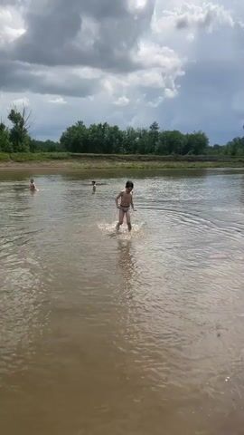 A group of young boys are playing in a shallow river. The water is murky brown and there are trees lining the opposite bank. The sky is cloudy. One boy in the foreground, wearing striped swim trunks, is splashing through the water towards the camera. Another boy, in the background, is further up the river. The overall scene depicts a casual, outdoor recreational activity.