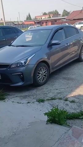 The video shows a gray Kia Rio sedan parked on a paved surface, possibly a driveway or parking lot. The car is viewed from the side and slightly from the front. The background includes other vehicles, buildings, and greenery. The lighting suggests it is daytime. There are no people visible in the video, and no actions are taking place other than the slight camera movement.