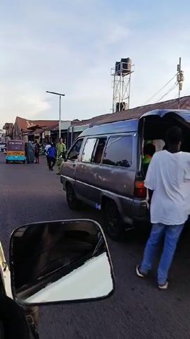 The video shows a street scene in what appears to be a West African city or town. The shot is taken from inside a vehicle. A light blue and brown auto rickshaw labeled "PTA-10288" is briefly in the foreground. People are walking along the side of the road and in the street. There are various shops and buildings lining the road. A man is pushing a wheelbarrow loaded with items. Several cars are parked along the road. There's a water tower visible in the background. A street light is also visible. The sky is overcast.