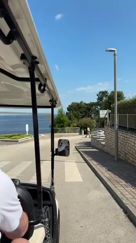 A golf cart drives along a paved road. The camera is positioned inside the golf cart, facing forward and slightly to the right. A person's shoulder and arm are visible in the lower left corner of the frame, indicating they are seated in the golf cart. The road has white crosswalk markings. To the left of the road is a body of water, likely the sea, with a clear blue sky above. Green vegetation and trees are visible on the right side of the road, along with a low stone wall and a signpost. The golf cart is moving forward, and the scene suggests a sunny day in a scenic location, possibly a resort or coastal area.