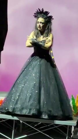 A woman in a black, glittery gown and a feathered headdress stands on a stage. She is singing and gesturing with her hands. The background is a soft pink. There are some colorful props to the right of the woman.
