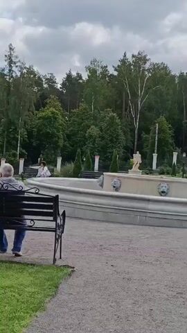 The video shows an outdoor scene in a park or garden. A large, ornate fountain with lion head spouts is visible in the center of the frame. Several people are walking around the fountain and in the surrounding area. There are benches, lampposts, and manicured trees and bushes. The sky is overcast. The camera appears to be handheld and is moving slightly. One person in the foreground is wearing a blue shirt and dark pants, and another person in the background is wearing a light blue dress. The audio consists of muffled conversation.