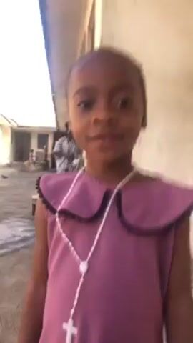 A young girl stands outside wearing a purple dress with a white rosary around her neck. She looks at the camera and smiles. A man can be heard talking in the background, and the girl puts her hand to her mouth while looking at the camera.  A building and a few other people are visible in the background.