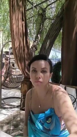 A woman with dark hair and makeup is sitting outside at a table, holding a glass of a light-colored liquid and drinking from it. She is wearing a blue and turquoise patterned dress. Behind her, there is a woven hanging chair and some large, brown curtains. Green foliage hangs overhead.