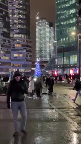 The video shows a city street at night, wet with puddles. People are walking across the street in both directions, many dressed in winter coats and hats. There is a Christmas tree lit up with blue lights in the middle ground. Tall buildings surround the street, with many windows lit up. A tall building in the background appears to be under construction with a crane visible at the top. The lighting is mainly from streetlights and building lights. The general atmosphere seems busy and festive.