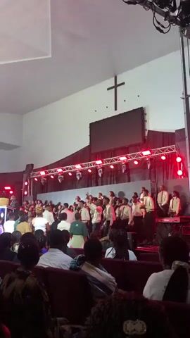 A large group of people, appearing to be a choir, are gathered on a stage in a church. They are wearing formal attire, mostly white shirts and ties for the men and various colored dresses or skirts for the women. Red lights illuminate the stage from above. A large screen is mounted on the wall behind the choir, and a wooden cross is visible above the screen. In front of the stage, an audience is seated in pews, facing the performers. Some members of the choir are singing and gesturing, and the overall atmosphere appears to be one of lively worship or performance. The church interior is spacious with white walls and a high ceiling.