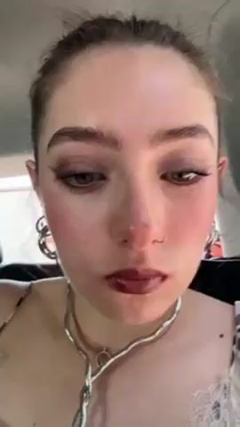 A close-up video features a young woman applying dark lipstick. She has fair skin, brown hair pulled back, visible eye makeup and blush, a nose ring, and multiple necklaces and earrings. The lipstick tube is black with a gold top and reads "300 Coffee Bean." Behind her, blurry figures of people are visible, suggesting she might be in a car.