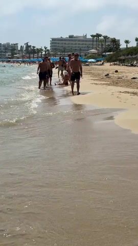 The video shows a group of men walking along a sandy beach next to the ocean. The water is clear and the waves are gently lapping at the shore. The men are wearing swim trunks and appear to be enjoying the warm weather. In the background, there are buildings that look like hotels or resorts, palm trees, and other beachgoers. The sky is blue and the sun is shining. The overall scene depicts a typical beach vacation.