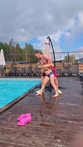 A young boy wearing blue swim trunks and goggles stands on the edge of a swimming pool. An adult male wearing red swim trunks and white Crocs holds the boy by his arms, pushing him into the pool. The boy falls into the water, creating a splash. The adult watches the boy as he swims in the pool. The scene takes place outdoors on a wooden deck next to the pool. There are lounge chairs and umbrellas in the background. Two pink flip-flops are visible on the deck in the foreground. The sky is cloudy.