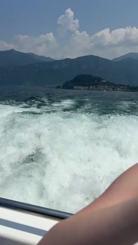 This is a first-person perspective video clip filmed from a boat on a body of water, likely a lake, on a sunny day. The camera is positioned to capture the churning white wake of the boat and the distant shoreline. In the background, there are lush green mountains that rise majestically from the water's edge. A small, picturesque town with terracotta roofs is nestled on a hillside overlooking the lake, possibly Griante, Italy. The sky is a clear blue with a few fluffy white clouds, suggesting fair weather. The sun appears to be high in the sky, indicating it is likely midday. The overall mood is one of travel and enjoyment, with the motion of the boat and the scenic beauty of the location contributing to a sense of relaxation and adventure. There is no visible text within the clip. The clip lasts for approximately 4 seconds.