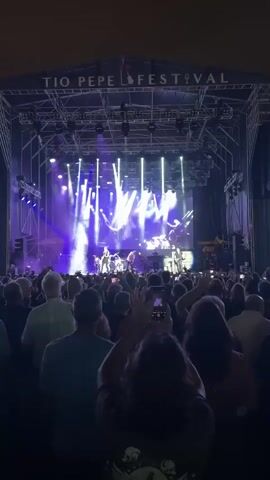 The video shows a concert performance by a band on a brightly lit stage. The stage has a large screen behind the band, displaying abstract visuals. Several band members are visible, including a guitarist on the left, a drummer in the center, a keyboardist further back, and another guitarist on the right. The audience is gathered in front of the stage, many of whom are holding up their phones to record the performance. The venue appears to be outdoors, with a large structure above the stage that supports lighting and sound equipment. The overall atmosphere is energetic, with the audience engaged and the band actively performing.