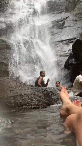 A group of people are at the base of a waterfall. Two people are partially submerged in the water near the base of the falls, while another person is sitting on a rock to the side. The camera angle is low, showing the feet of another person in the foreground, likely the one taking the video. The waterfall is a multi-tiered cascade over dark, rocky terrain. The scene is generally bright and natural, with the focus on the waterfall and the people enjoying the water.