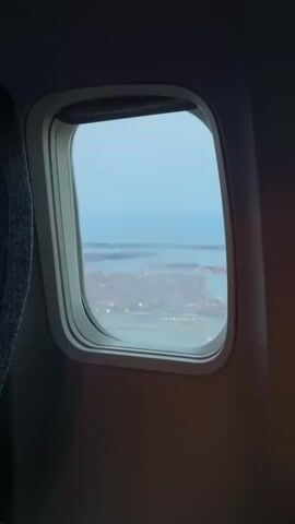The video shows a view from an airplane window. Outside the window, a city can be seen below, with water and buildings illuminated by lights. The sky is a pale blue, suggesting it is either dawn or dusk. The camera is positioned inside the airplane, looking out. The window frame and a bit of the airplane seat are visible in the foreground. The footage is shaky, indicating movement, likely from the airplane in flight or during landing/takeoff.