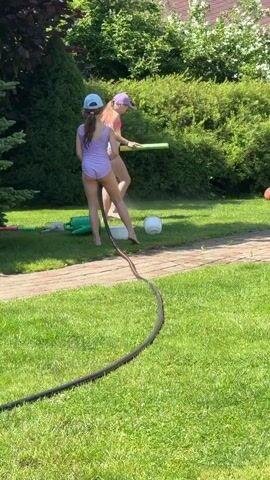 Two young girls are playing with a water hose in a grassy backyard on a sunny day. The girl on the left wears a purple swimsuit and a blue baseball cap. The girl on the right wears a multi-colored swimsuit and a purple baseball cap. They are both barefoot. The girl on the right is holding a green and yellow water squirter. There is a white ball on the ground near the girls, and a green watering can and other toys are visible in the background. A black garden hose snakes across the grass. The area appears to be a suburban backyard with mature trees and bushes. The girls are interacting with the water hose, possibly spraying each other or playing a game. The overall scene depicts a playful summer activity.
