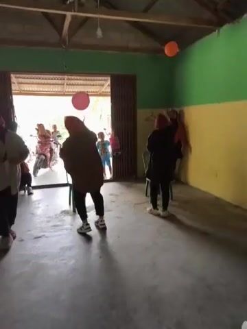 The video shows an indoor space that appears to be a community hall or large room with an open doorway leading outside. The walls are painted in green and yellow. The ceiling is made of corrugated metal with visible wooden beams. Several people are present inside the room, and some are visible through the open doorway. In the foreground, a person wearing a brown hooded jacket and dark pants is facing away from the camera, appearing to be dancing or performing an action. Two green plastic chairs are visible in the room. In the background, through the doorway, several people are gathered around motorcycles. Two balloons, one pink and one orange, are hanging from the ceiling. The floor is concrete. The overall atmosphere suggests a gathering or event.