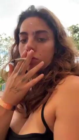 A woman with long brown hair and an orange wristband is holding a lit cigarette in her left hand and exhaling smoke directly into the camera. The woman is wearing a black top. The background shows trees and other people. The woman's left hand is visible, with the word "feel" tattooed on it. The wristband says "ROOTS."