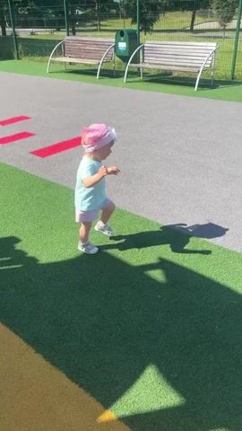 A young child wearing a pink hat and a light blue t-shirt is on a playground. The child is playing with their shadow, first reaching towards it, then bending down to touch it. The playground has a grey surface with green patches and red markings. In the background, there are benches and a fence.