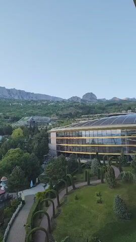 The video shows a scenic view of a mountainous landscape with buildings and lush greenery. The camera pans slowly from right to left, revealing a large, modern hotel with multiple stories and many windows. In the foreground, there is a beautifully landscaped garden with a winding pathway and neatly trimmed shrubs. Beyond the hotel and garden, the view opens up to a landscape densely covered with trees and scattered buildings. In the distance, the horizon is dominated by a mountain range under a clear, light blue sky.