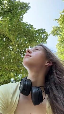 The video shows a young woman from the chest up, looking upwards at an angle. She has long, dark brown hair, some of which is blowing across her face. She is wearing a light yellow top and black headphones are around her neck. The background is filled with green leaves and blue sky, suggesting an outdoor setting, possibly a park or forest. Her expression changes from a neutral gaze to a slight smile as she looks towards the camera.